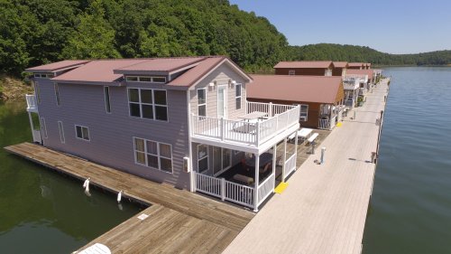 Aerial view of houseboats on a calm lake, surrounded by green hills under a clear blue sky.