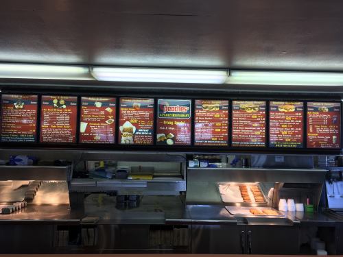 Menu board at a fast food restaurant featuring various food items and prices, with a kitchen area visible below.