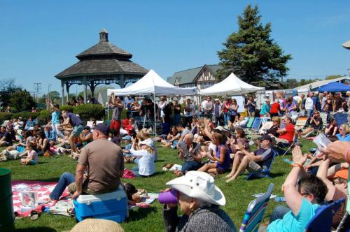 A lively outdoor event with a large crowd seated on the grass, enjoying performances near a gazebo and tents.