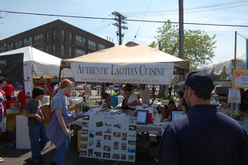 A bustling outdoor market stall featuring "Authentic Laotian Cuisine" with people enjoying food and activities.