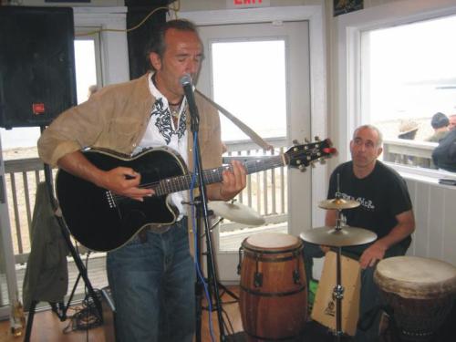 A musician sings and plays guitar while a drummer accompanies him in a beachside venue.
