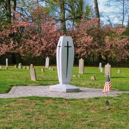 A memorial with two crosses stands in a cemetery, surrounded by gravestones and blooming trees, with an American flag nearby.