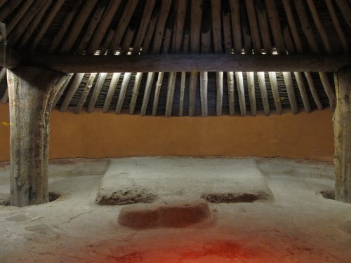 Interior of a traditional structure with a thatched roof, featuring wooden beams and a clay floor. Soft lighting illuminates the space.