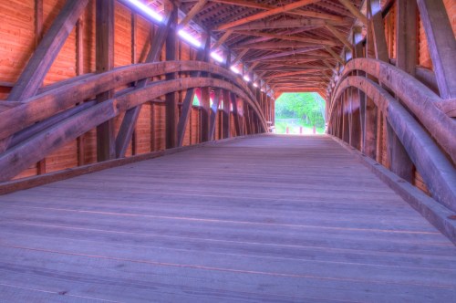 A wooden covered bridge interior with arched beams and a wooden floor, leading to a bright exit.