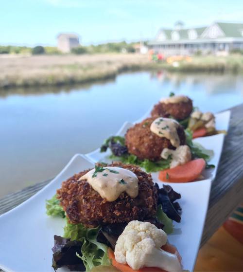 Three golden-brown croquettes topped with sauce, served on a bed of greens and vegetables by a scenic water view.