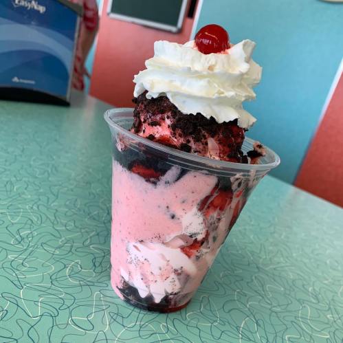 A colorful dessert in a cup featuring layers of pink ice cream, chocolate crumbs, whipped cream, and a cherry on top.