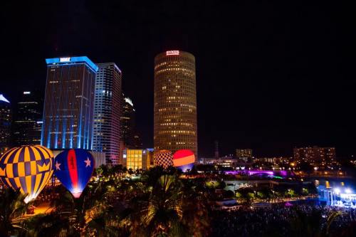Colorful hot air balloons illuminate the night sky above a vibrant city skyline with tall buildings and a lively crowd below.