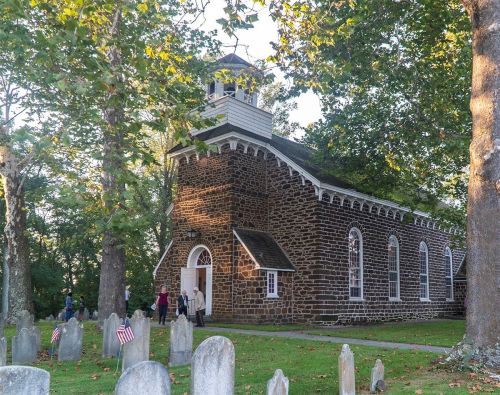 Historic stone church surrounded by trees and a graveyard, with people walking nearby and small flags on gravestones.