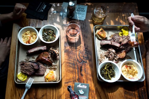 Two trays of barbecue with sides, including mac and cheese and greens, on a wooden table with drinks and a menu.