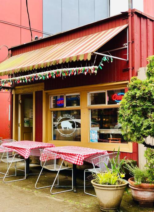 A cozy restaurant exterior with red and white checkered tables, a striped awning, and colorful lights.