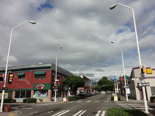 A street view of a small town with shops, traffic lights, and cloudy skies overhead.