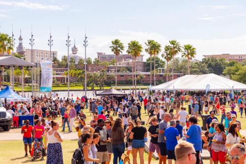 A lively outdoor festival with a large crowd, palm trees, and tents by a waterfront under a clear blue sky.