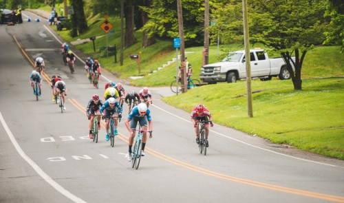A group of cyclists racing down a tree-lined road, with spectators and a parked truck in the background.