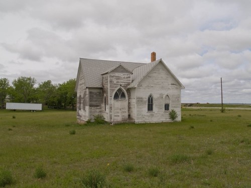 Abandoned white church with arched windows, surrounded by overgrown grass and a cloudy sky in the background.