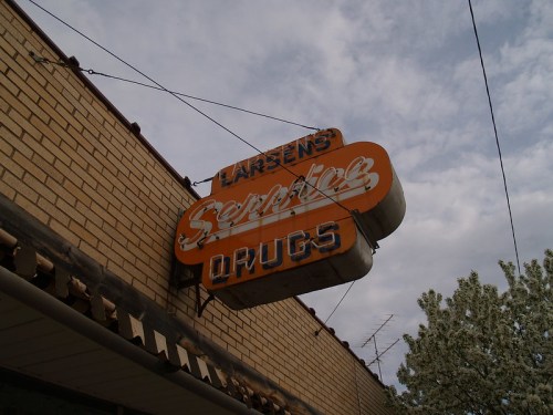 Vintage sign for "Larsen's Service Drugs" hanging from a brick building under a cloudy sky.