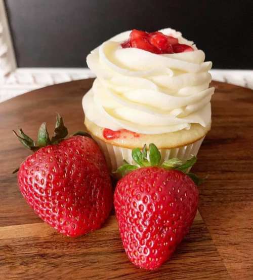 A vanilla cupcake topped with swirled frosting and strawberry topping, accompanied by two fresh strawberries on a wooden surface.