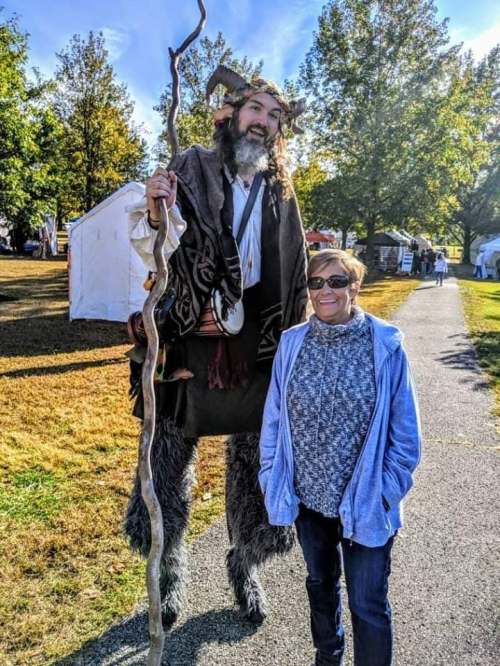 A tall, costumed figure with a staff stands next to a smiling woman in a park, surrounded by trees and tents.