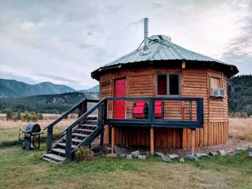 A round wooden cabin with a metal roof, red door, and deck, surrounded by mountains and grassy fields.