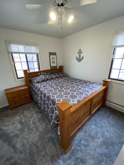 Cozy bedroom featuring a wooden bed, gray patterned bedding, nightstand, and windows with natural light.