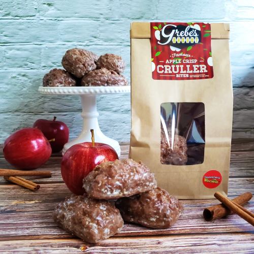 A bag of Grebe's Apple Crisp Cruller Bites with crullers and fresh apples on a wooden table, accented by cinnamon sticks.