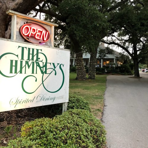 Sign for "The Chimneys" with neon "Open" light, surrounded by trees and festive lights, leading to a dining establishment.