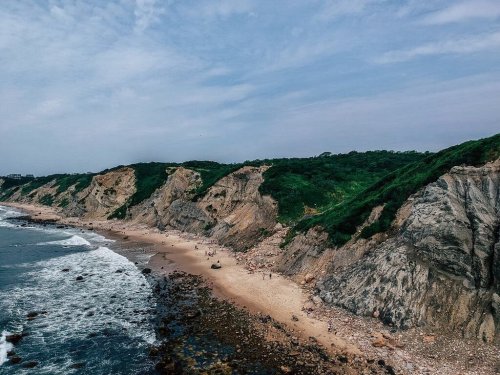 A scenic beach with rocky cliffs and lush greenery under a cloudy sky, with people walking along the shore.