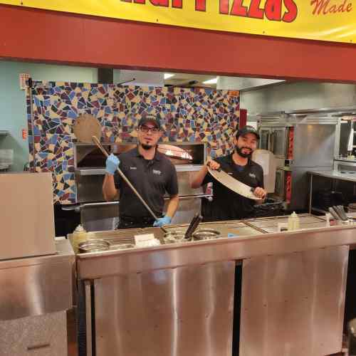 Two chefs in a pizza kitchen, one holding a pizza peel and the other a large knife, smiling at the camera.