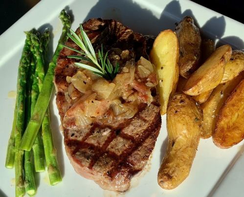 A grilled steak topped with onions, served with roasted potatoes and asparagus on a white plate.