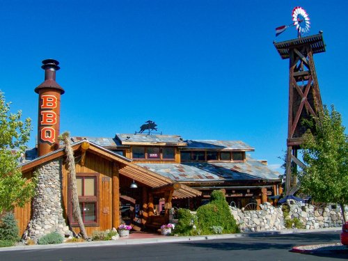 A rustic BBQ restaurant with a stone chimney, wooden exterior, and a windmill nearby under a clear blue sky.