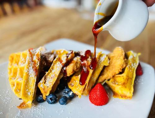 A plate of waffles topped with fried chicken, blueberries, strawberries, and syrup being poured over the dish.