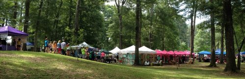 A vibrant outdoor market with colorful tents set among trees, featuring people browsing and enjoying the event.