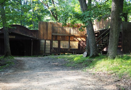 A rustic wooden building with multiple levels, surrounded by trees and a dirt path leading up to it.