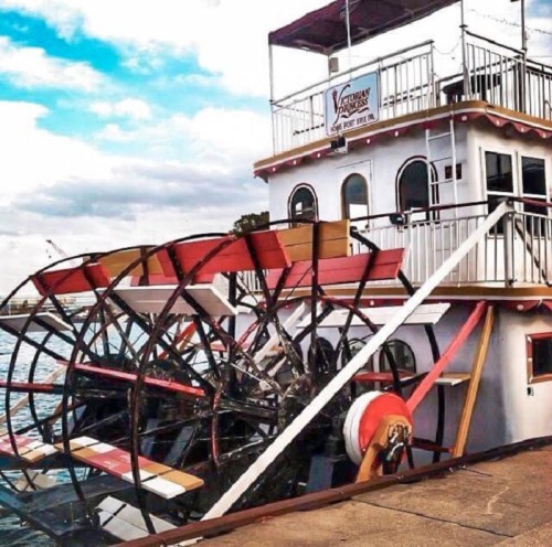 A riverboat with a large paddle wheel docked by the water, featuring a multi-level deck and vibrant colors.