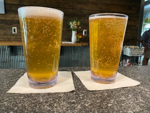 Two glasses of bubbly golden beer on a granite countertop, resting on white napkins. Background features a rustic wooden wall.