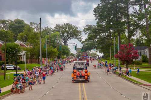 A lively street scene with a parade, featuring crowds of people and colorful floats under a cloudy sky.