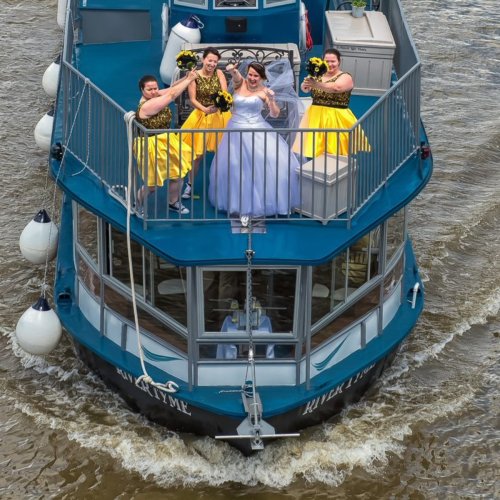 A bride in a white dress celebrates on a boat with three bridesmaids in yellow dresses, surrounded by water.