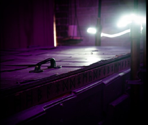 A close-up of a dark, textured surface with mysterious symbols, illuminated by purple light in a dimly lit space.