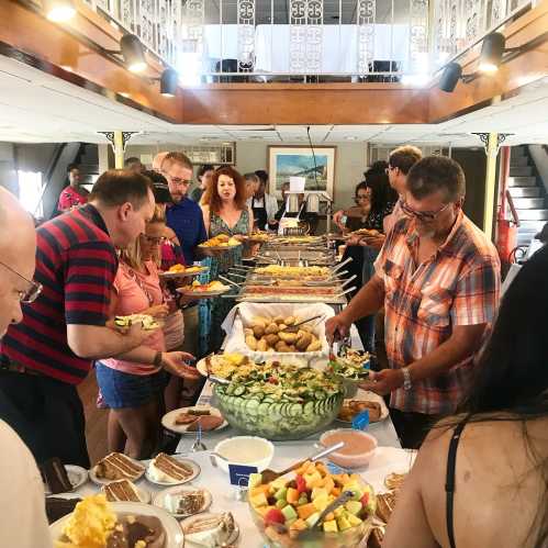 A diverse group of people enjoying a buffet with various dishes, fruits, and desserts in a spacious dining area.