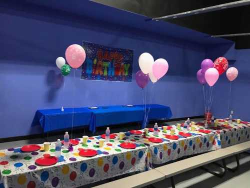 A festive birthday party setup with colorful balloons, tablecloths, and plates on a blue wall background.