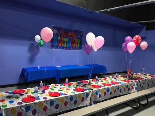 A festive birthday party setup with colorful balloons, tablecloths, and plates on a blue wall background.