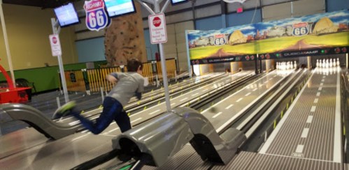 A child bowling in an indoor alley, with colorful signs and a rock climbing wall in the background.