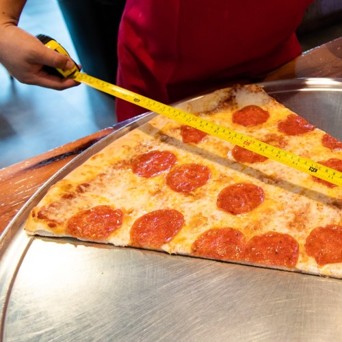A person measures a large slice of pepperoni pizza with a tape measure on a metal tray.