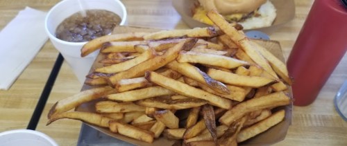 A large serving of golden-brown French fries in a tray, with a drink and a burger in the background.