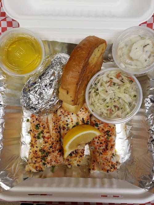 A takeout container with seasoned fish, a lemon wedge, a baked potato, coleslaw, garlic bread, and dipping sauce.