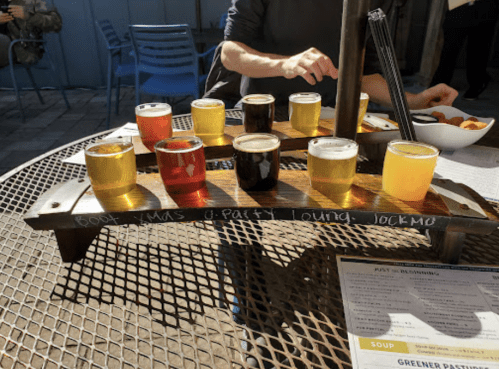 A wooden flight tray with eight small glasses of various beers on a table, with a menu and snacks in the background.