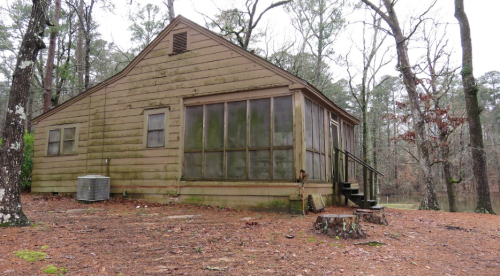 A weathered wooden cabin surrounded by trees, with a screened porch and steps leading up to the entrance.
