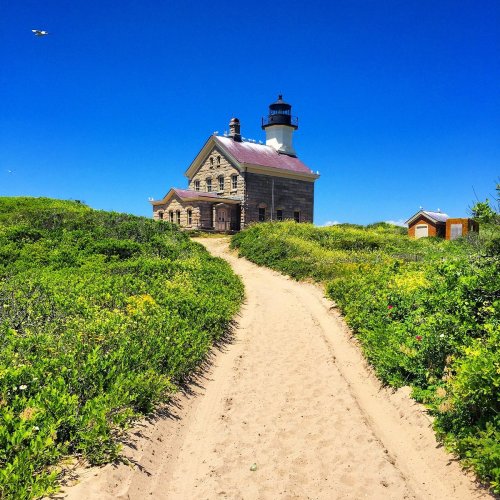 A sandy path leads to a historic lighthouse surrounded by lush greenery under a clear blue sky.