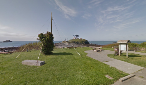 A coastal scene featuring grassy areas, a picnic table, and a distant lighthouse on a rocky island under a clear sky.