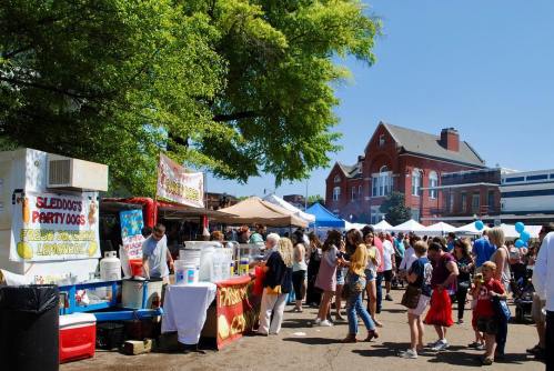 A bustling outdoor market with food stalls, people browsing, and a historic building in the background under a clear blue sky.