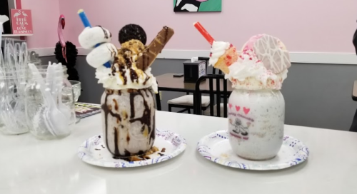 Two colorful milkshakes in jars, topped with whipped cream, cookies, and candy, served on paper plates.